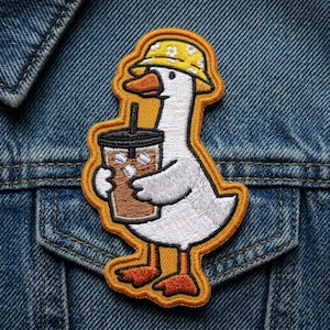 May include: Embroidered patch featuring a white goose wearing a yellow floral bucket hat and holding an iced coffee. The patch has an orange border and is attached to a denim jacket. The goose has orange feet and a beak.