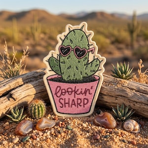 May include: An embroidered patch featuring a green cactus wearing pink heart-shaped sunglasses, planted in a pink pot with the text "lookin' SHARP" stitched in dark pink thread. The patch has a cream-colored border and a die-cut shape.