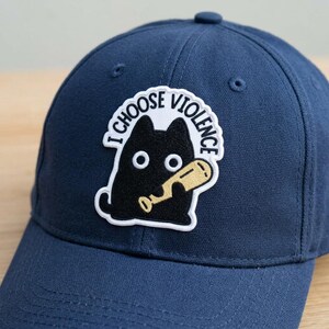 May include: A navy blue baseball cap featuring a patch with the text "I Choose Violence." The patch depicts a black cat holding a baseball bat. The cap has a curved brim and a button on top.