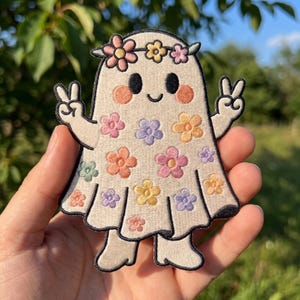May include: An embroidered patch featuring a cream-colored ghost character with a black outline. The ghost wears a floral crown and has colorful embroidered flowers across its body. It has black eyes, a small smile, pink cheeks, and both hands raised in a peace sign gesture.