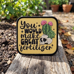 May include: A square embroidered patch with a yellow background and a black border. The text reads "You WOULD make GREAT fertilizer" in black lettering. An illustration of a human skull with a green cactus and pink flowers is included.
