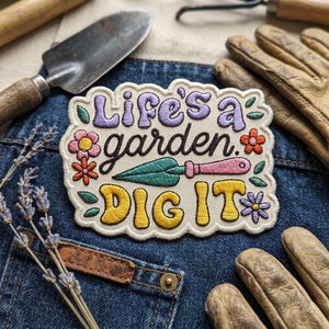 May include: An embroidered patch with a cream background featuring the text Life's a garden in purple and dark brown, and DIG IT in yellow. The design includes a green trowel with a pink handle, small orange, pink, and purple flowers, and green leaves.