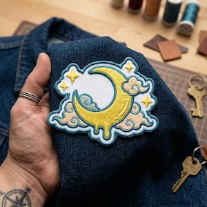 May include: Embroidered patch featuring a yellow crescent moon with a white center, surrounded by yellow stars and peach-colored clouds. The patch has a light blue border and is displayed on a dark blue denim fabric.