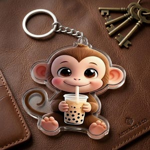 May include: A brown monkey keychain with a clear plastic border. The monkey is holding a cup of boba tea with a straw. The keychain has a silver chain and ring. The background is a brown leather surface.