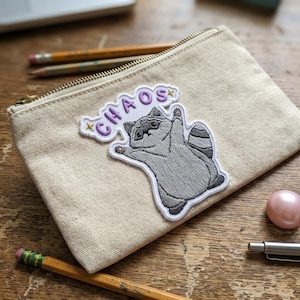 May include: A beige canvas pencil case with a gold zipper, featuring an embroidered patch of a gray raccoon with the word "CHAOS" in purple. The raccoon is raising one arm. A pink sphere and pencils are in the background.