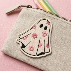 May include: A beige pencil case with a zipper, featuring an embroidered ghost patch. The ghost is white with pink flower accents and black eyes. Pencils in pink, yellow, and green are visible.