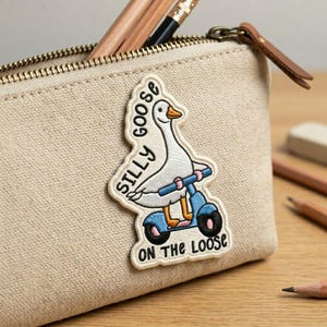 May include: Beige canvas pencil case with a zipper closure and a brown leather pull tab. The case features an embroidered patch of a white goose riding a blue scooter, with the text "SILLY GOOSE ON THE LOOSE". Pencils are visible inside.