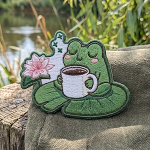 May include: An embroidered patch featuring a green frog with closed eyes sitting on a lily pad, holding a white mug filled with dark brown liquid. A pink water lily flower is positioned to the left, with small green cross-stitch details nearby.