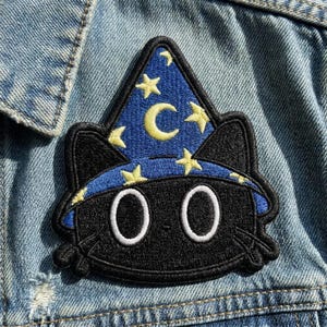 May include: An embroidered patch featuring a black cat wearing a blue wizard hat decorated with yellow stars and a crescent moon. The cat has large white eyes and black whiskers, with the entire design outlined in a thick black border.