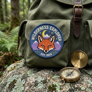 May include: A green canvas backpack with a "Wilderness Explorer" patch featuring a fox, mountains, and a crescent moon. A brass compass sits on a rock. The patch reads "Est. 2023".