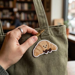 May include: An olive green canvas tote bag with a brown embroidered croissant patch. A gold fountain pen is held near the bag's seam. The patch features a smiling croissant with brown feet and a white border.