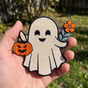 May include: An embroidered patch featuring a cream-colored ghost with a black outline. The ghost has a smiling face and holds an orange jack-o'-lantern bucket in one hand and a single orange flower with teal leaves in the other hand.