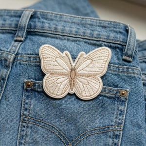 May include: A beige embroidered butterfly patch is affixed to the back pocket of a pair of blue denim jeans. The butterfly has detailed wing patterns and a cream-colored border. The jeans have classic stitching and metal rivets.