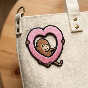 May include: A canvas tote bag with a decorative patch. The patch features a brown monkey inside a pink heart-shaped frame. The bag has a handle and a metal ring attached to the side.