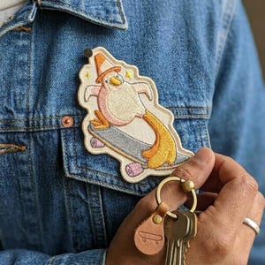 May include: Embroidered patch featuring a cartoon character riding a skateboard. The character has a cone hat, pink body, and yellow tail. The patch is attached to a denim jacket. A leather keychain with a skateboard design and keys are in the foreground.