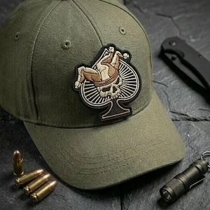 May include: Olive green baseball cap with a patch featuring a skull and jester design. A black folding knife, a small flashlight, and several bullets are also visible.