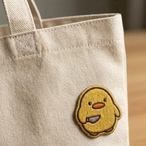 May include: A beige canvas tote bag with a yellow embroidered patch of a cartoon chick holding a knife. The chick has an orange beak and brown outline. The patch is sewn onto the front of the bag.