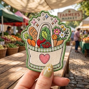 May include: An embroidered patch featuring a cream-colored tote bag with a pink heart, filled with a baguette, carrots, a red tomato, strawberries, and a bouquet of colorful flowers. The design includes green gingham trim and small sparkling stars.