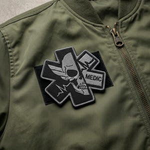 May include: Olive green bomber jacket with a black and gray Medic patch. The patch features a skull design within a cross, with the word "MEDIC" visible. The jacket has a zipper closure.