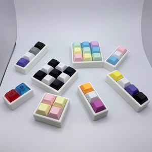 May include: A collection of colorful keyboard keycaps in various arrangements. The keycaps are in white rectangular trays, with colors including black, white, gray, purple, pink, blue, yellow, and orange. The image is taken on a white background.