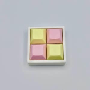 Pode incluir: Um prato quadrado de cerâmica branca contém quatro teclas de teclado quadradas em cores pastel. Duas são amarelas e duas rosas. As teclas estão dispostas numa grelha de dois por dois. O fundo é cinzento claro liso.