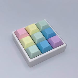 Könnte beinhalten: Ein quadratisches, weißes Tablett mit neun quadratischen Objekten in Pastellfarben. Die Farben sind Rosa, Gelb, Grün, Blau und Lila. Die Objekte sehen aus wie Tasten und sind wahrscheinlich dekorativ oder funktional.
