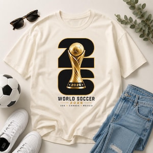 以下が含まれることがあります： 2026年ワールドサッカーの金色のトロフィーデザインが施されたクリーム色のTシャツ。「WORLD SOCCER 2026 USA CANADA MEXICO」の文字入り。青いジーンズ、白いスニーカー、サングラスとコーディネートされています。