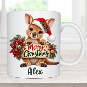 May include: A white ceramic mug featuring a cartoon kangaroo wearing a red and white Santa hat. The kangaroo holds a wooden sign that reads Merry Christmas in red and green text. A plaid bow and holly berries decorate the sign. The name Alex is printed below.