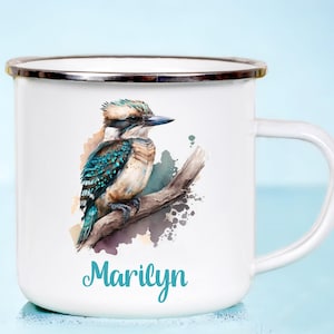 May include: White enamel mug with a watercolour illustration of a kookaburra. The bird has blue, teal, and brown feathers. The name "Marilyn" is written in teal script below the bird. The mug has a silver rim.