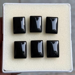 Puede incluir: Seis piedras preciosas negras rectangulares y pulidas están dispuestas en una caja blanca. Cada piedra tiene una superficie brillante que refleja la luz. Las piedras están espaciadas uniformemente, lo que sugiere un conjunto o colección de artículos similares.