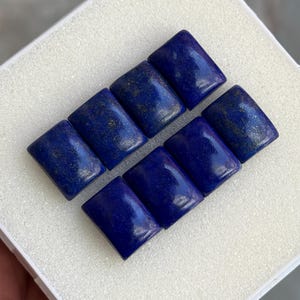 May include: Eight rectangular lapis lazuli gemstones, each with a deep blue hue and flecks of gold. The stones are arranged in two rows inside a white box, showcasing their polished surfaces and natural variations.