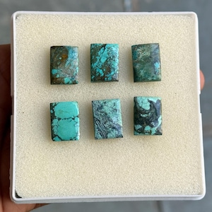 May include: Six rectangular turquoise gemstones with varying shades of blue and green are displayed in a white tray. The stones have unique patterns and textures, showcasing natural variations. The gemstones are likely for jewelry making.