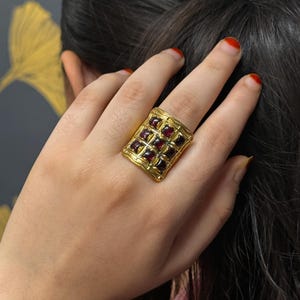 Puede incluir: Un anillo dorado con una cara rectangular, con un patr&oacute;n de rejilla y piedras preciosas cuadradas de color rojo oscuro. El anillo se lleva en una mano con esmalte de u&ntilde;as rojo, sobre un fondo de pelo oscuro y un dise&ntilde;o de hoja dorada.
