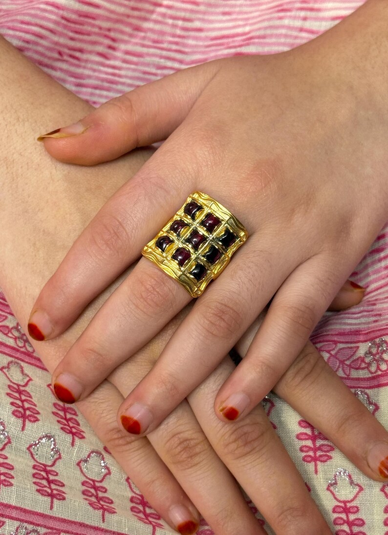 Puede incluir: Un anillo dorado con una cara cuadrada, engastado con una cuadr&iacute;cula de piedras preciosas de color rojo oscuro. El anillo est&aacute; en una mano con esmalte de u&ntilde;as rojo. El dise&ntilde;o del anillo presenta un patr&oacute;n texturizado en forma de gofre.