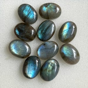 Könnte beinhalten: Eine Ansammlung von neun ovalen Labradorit-Edelsteinen, die jeweils eine glatte, polierte Oberfläche aufweisen, mit irisierenden blauen, grünen und goldenen Lichtreflexen aus dem Inneren der dunkelgrauen, halbtransparenten Steine.