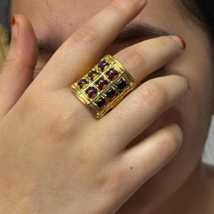 Puede incluir: Un anillo dorado con una cara rectangular, con una cuadr&iacute;cula de piedras preciosas cuadradas de color rojo oscuro. El anillo tiene un dise&ntilde;o ornamentado y texturizado alrededor de las piedras preciosas. El anillo se lleva en un dedo.