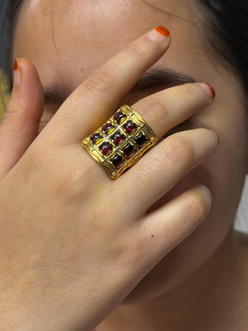 Puede incluir: Un anillo dorado con una cara rectangular, con una cuadr&iacute;cula de piedras preciosas cuadradas de color rojo oscuro. El anillo tiene un dise&ntilde;o ornamentado y texturizado alrededor de las piedras preciosas. El anillo se lleva en un dedo.