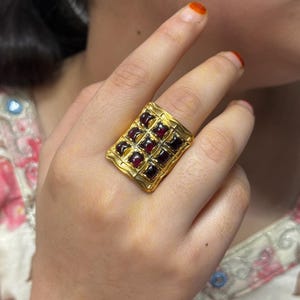 Puede incluir: Un anillo dorado con una cara rectangular adornada con una cuadr&iacute;cula de piedras preciosas cuadradas de color rojo intenso. El anillo presenta un borde texturizado, posiblemente martillado, que sugiere un estilo vintage o artesanal. Las piedras est&aacute;n espaciadas uniformemente.