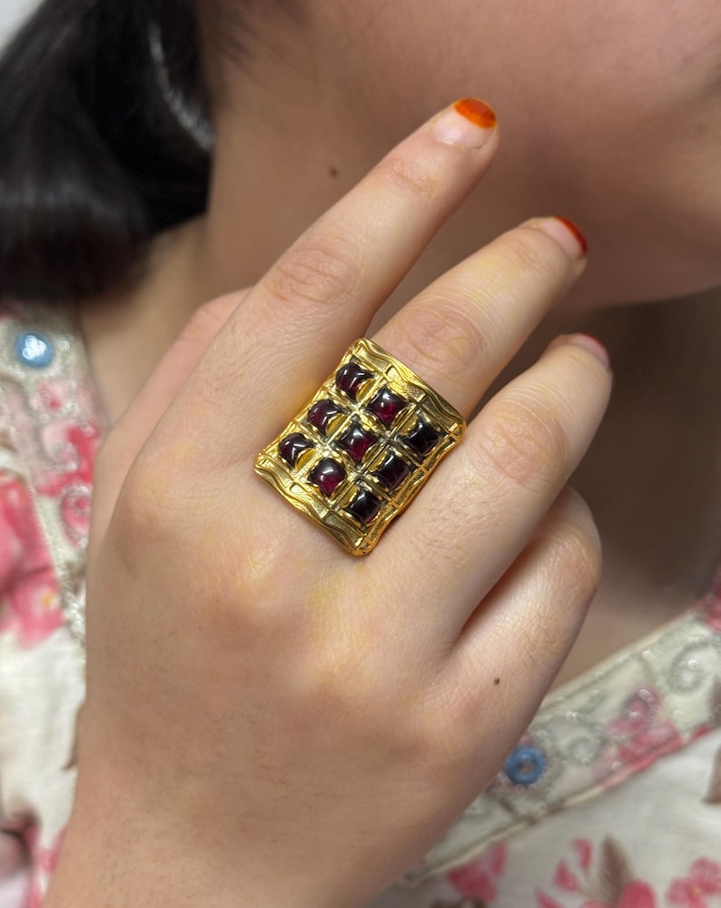 Puede incluir: Un anillo dorado con una cara rectangular adornada con una cuadr&iacute;cula de piedras preciosas cuadradas de color rojo intenso. El anillo presenta un borde texturizado, posiblemente martillado, que sugiere un estilo vintage o artesanal. Las piedras est&aacute;n espaciadas uniformemente.