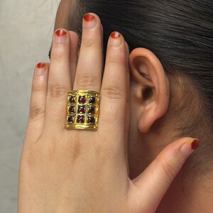 Puede incluir: Un anillo dorado con una cara rectangular. El anillo est&aacute; engastado con m&uacute;ltiples piedras preciosas rojas oscuras de corte cuadrado. El dise&ntilde;o del anillo presenta un patr&oacute;n de rejilla y las piedras preciosas est&aacute;n espaciadas uniformemente.