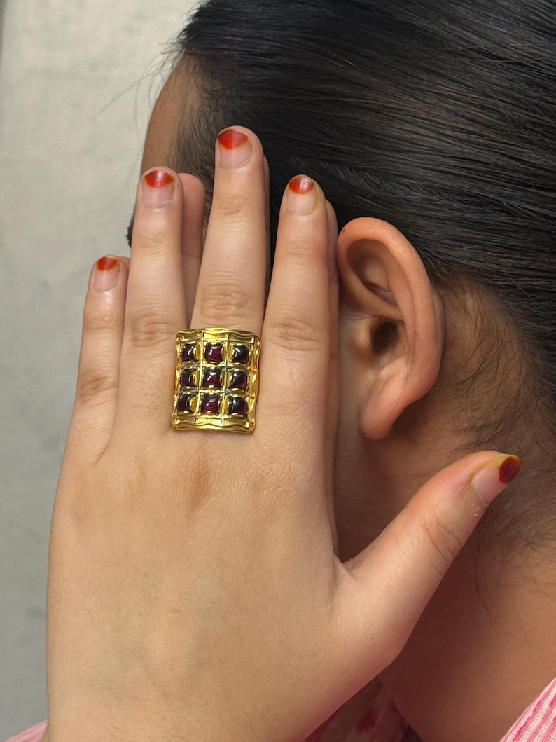 Puede incluir: Un anillo dorado con una cara rectangular. El anillo est&aacute; engastado con m&uacute;ltiples piedras preciosas rojas oscuras de corte cuadrado. El dise&ntilde;o del anillo presenta un patr&oacute;n de rejilla y las piedras preciosas est&aacute;n espaciadas uniformemente.