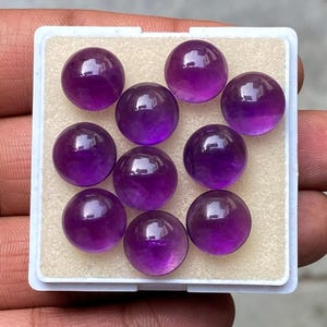 May include: Ten polished, round amethyst gemstones displayed in a white plastic tray. The gemstones are a deep purple color with a smooth, reflective surface. The tray is held by a hand.