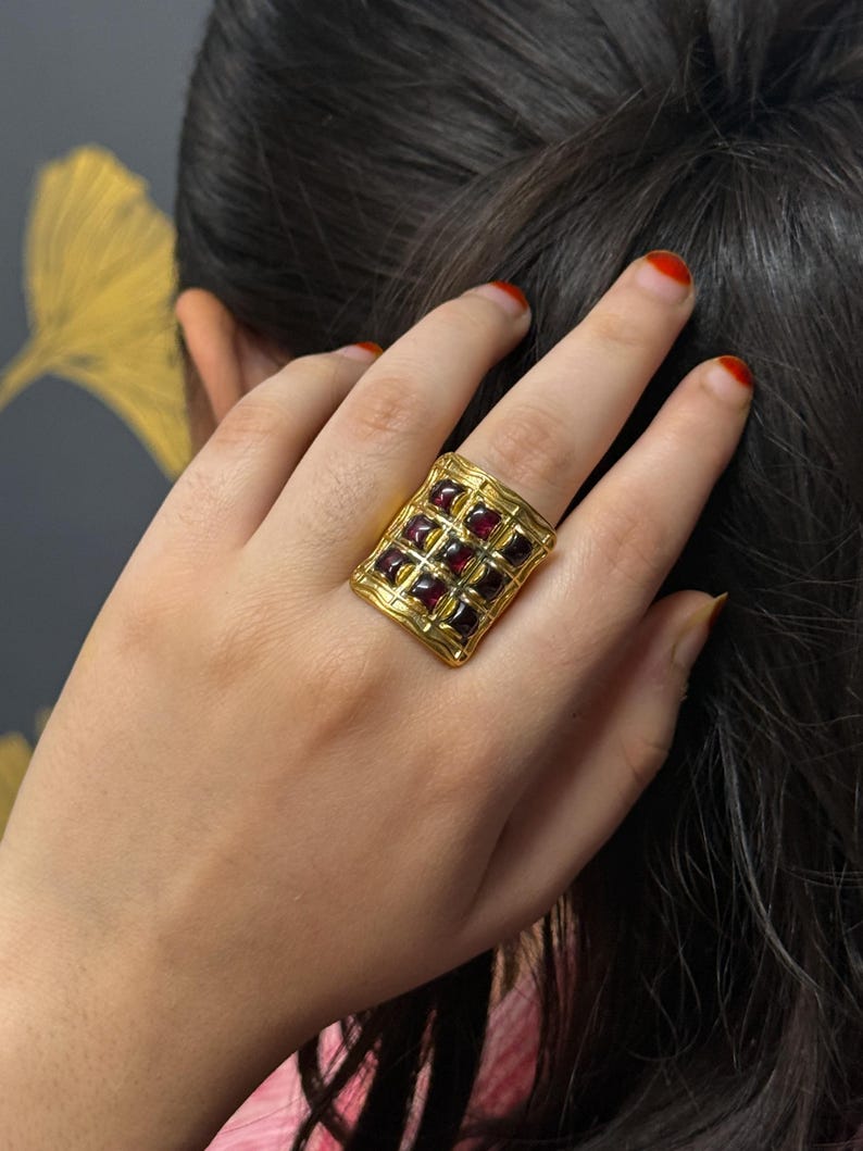 Puede incluir: Un anillo dorado con una cara rectangular, con un patr&oacute;n de rejilla y piedras preciosas cuadradas de color rojo oscuro. El anillo se lleva en una mano con esmalte de u&ntilde;as rojo. El fondo es una pared gris oscuro con un dise&ntilde;o de hoja dorada.