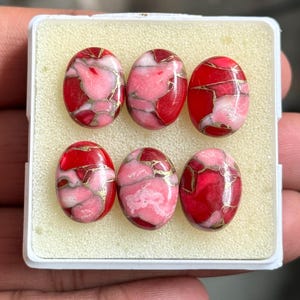 Può includere: Sei pietre preziose ovali in un vassoio bianco. Le pietre presentano un mix di rosso, rosa e oro. Le pietre sono lucide e brillanti. Il vassoio è posto su uno sfondo neutro. Adatto per la creazione di gioielli.