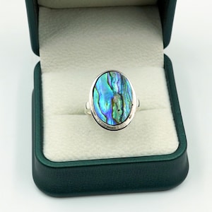 May include: An oval-shaped ring featuring a polished abalone shell inlay with iridescent blue, green, and purple hues. The shell is set within a smooth, polished silver-toned metal band, displayed inside a dark green hinged jewelry box.