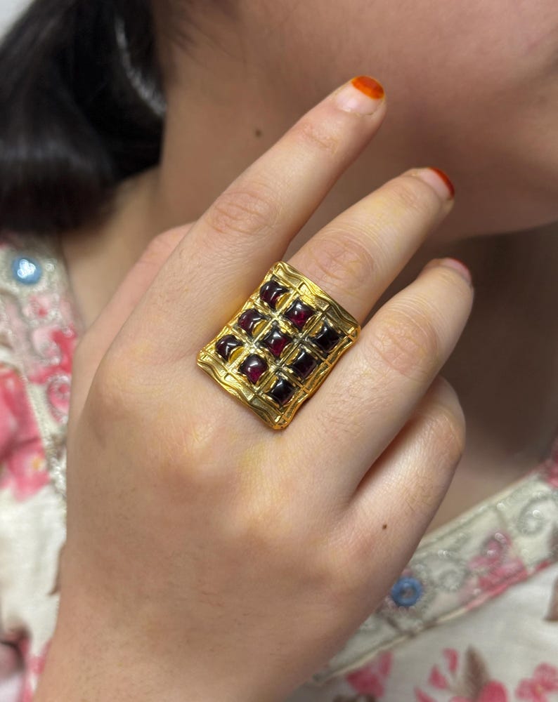 Puede incluir: Un anillo dorado con una cara rectangular, engastado con una cuadr&iacute;cula de piedras preciosas cuadradas de color rojo intenso. El anillo tiene un dise&ntilde;o texturizado, posiblemente martillado, en los bordes, lo que le da un aspecto vintage o artesanal. Las piedras preciosas est&aacute;n espaciadas uniformemente.