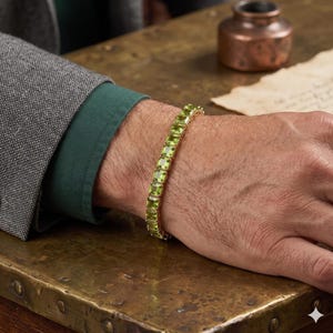May include: A gold bracelet featuring rectangular, light green gemstones. The bracelet is worn on a wrist, with a green shirt and gray blazer in the background. The gemstones are approximately 0.25 inches in length.