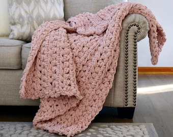 May include: A chunky knit throw blanket in a soft dusty pink color featuring a thick, textured braided pattern. The blanket is draped over the arm of a light gray upholstered sofa with visible nailhead trim along the front edge of the armrest.