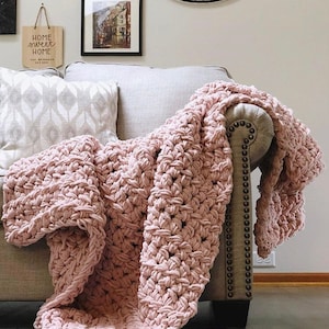 May include: A soft, pink crocheted throw blanket draped over a light gray couch. The blanket has a textured, chunky stitch pattern. The couch has a tufted back and a wooden frame.