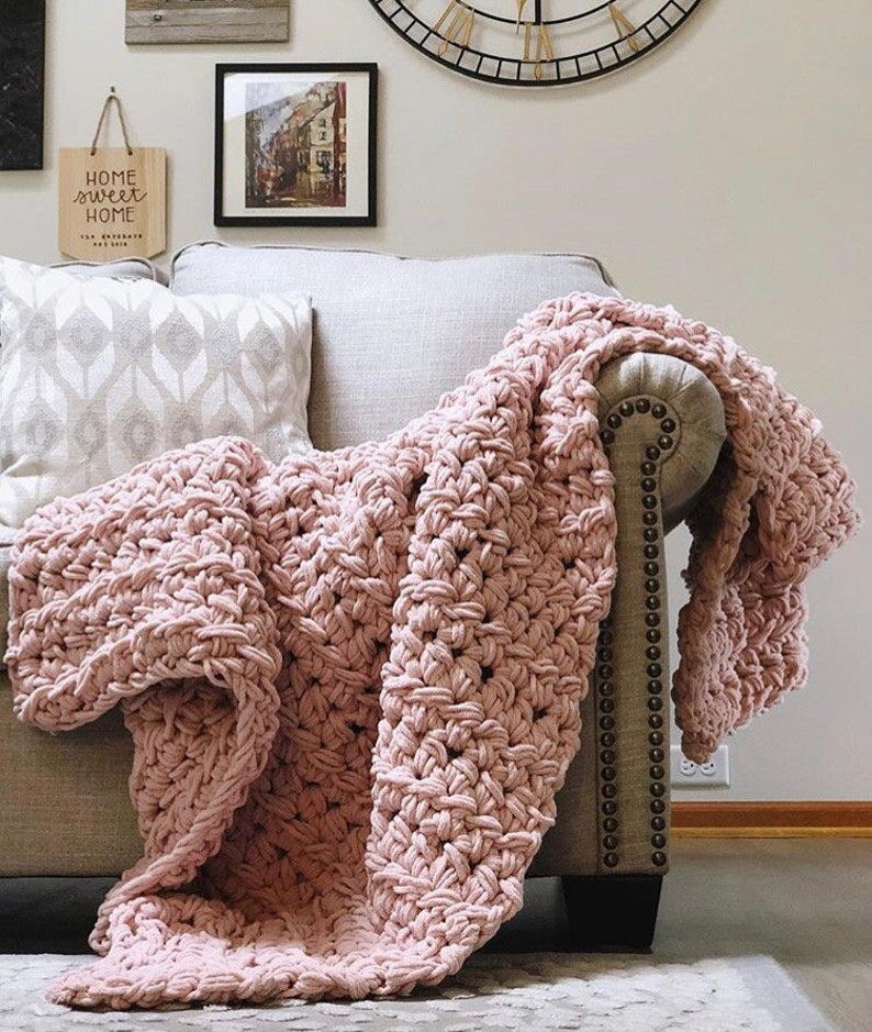 May include: A soft, pink crocheted throw blanket draped over a light gray couch. The blanket has a textured, chunky stitch pattern. The couch has a tufted back and a wooden frame.